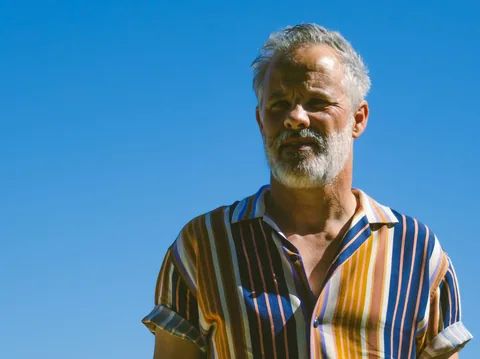 Illustrative photograph of a man with a grey beard in a striped shirt under a blue sky